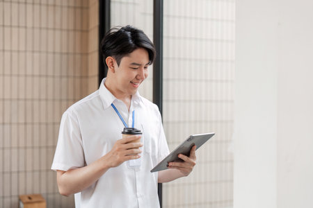 Young Asian male office worker standing and smiling while using tablet in modern workspace, enjoying...