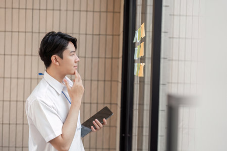 Young Asian male office worker contemplating sticky notes on glass wall in modern workspace, focusing on planning and strategy.の写真素材