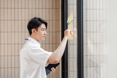 Young Asian male office worker organizing sticky notes on glass wall in modern workspace, focusing...