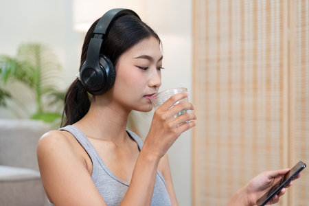 Hydration and Tech. A young woman sipping water while engaging with her smartphone and enjoying music.の写真素材