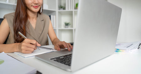 Organized Workspace and Productivity. A woman writes notes while using her laptop in a clean office...