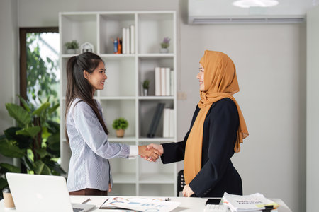 Diverse businesswomen shaking hands in a modern office, symbolizing partnership and collaboration.の写真素材