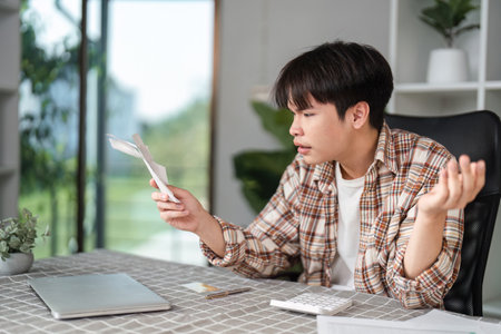 Young adult frustrated with financial bills while calculating expenses at home.の写真素材