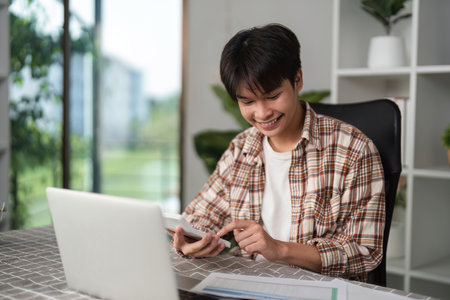 Young man using smartphone while reviewing financial statements at home.の写真素材