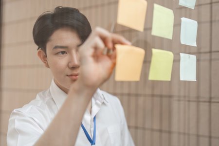 Young professional arranging sticky notes on glass wall, organizing ideas and enhancing workflow in modern office.の写真素材