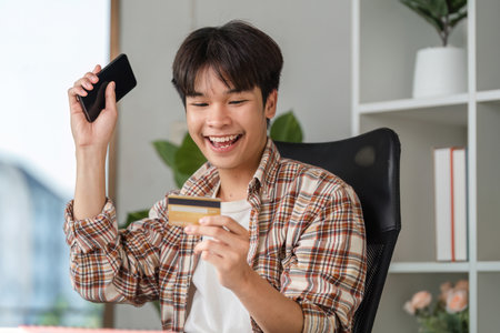 Joyful young man celebrating successful financial management with smartphone and credit card.の写真素材