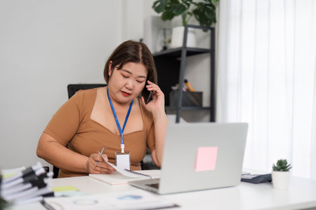 Young Female Office Worker Engaged in Phone Call and Taking Notes at Modern Workplace Deskの写真素材