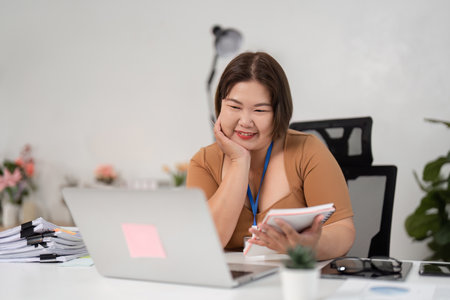 Happy Young Woman Analyzing Financial Documents at Home Office with Laptop and Notepadの写真素材