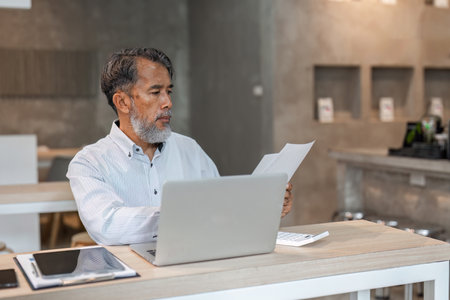 Diligent male businessman examining paperwork while working at a desk with laptop in a modern officeの写真素材