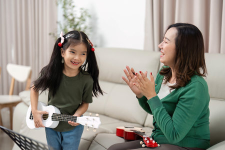 Learning and Fun. Young girl joyfully playing ukulele while mother claps in encouragement.の写真素材