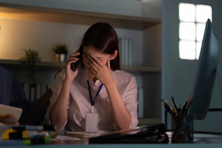 Stress and Overwhelm. Young woman on phone, feeling stressed at work in a dimly lit office.の写真素材