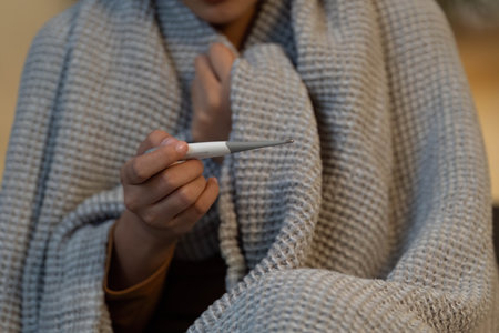 Health and Wellness. Woman checking temperature with thermometer while wrapped in blanket.
