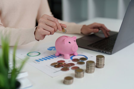 Savings Strategy. Woman depositing coins into piggy bank while working on laptop.の写真素材