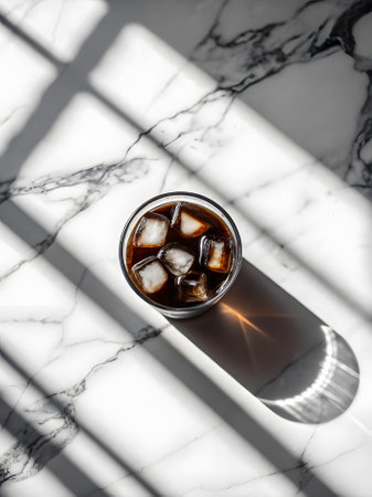 Top view of iced coffee with ice cubes in a glass on a marble surface, natural light casting...