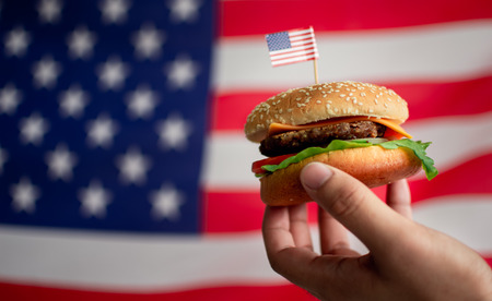 Man's hand holding a hamburger with the background of American flag for rememberance of Independence...