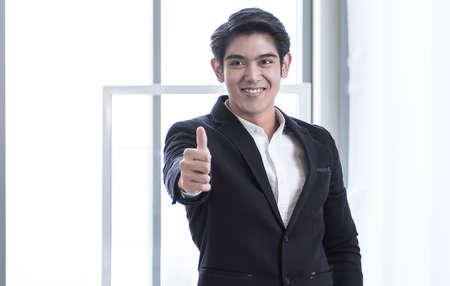 An Asian young business man wearing black formal black suit is successfully smiling and making thumb upの写真素材
