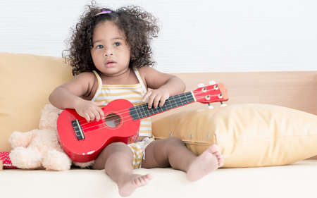Little african black girl with afro hair playing ukulele in living room at home.の写真素材