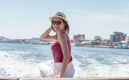Asian beautiful woman wearing hat and sunglasses, waving her hand while sitting on boat with background of beautiful landscape. Lifestyle and Travel Concept.の写真素材