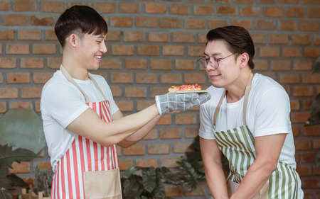 Asian handsome man smiling  giving a pizza to his boyfriend while cooking in the kitchen at home....
