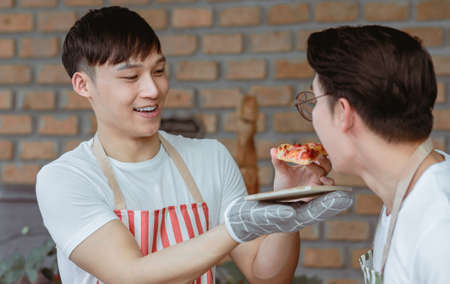 Asian handsome man smiling  giving a pizza to his boyfriend while cooking in the kitchen at home....