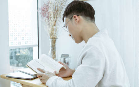 An asian man wearing eyeglasses and reading books beside window at home. Lifestyle and Leisure...