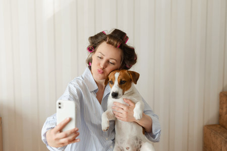 Beautiful young woman wearing pajamas with pink hair curlers holds a Jack Russell Terrier dog and takes a selfie on a smartphone at home on a sunny morningの写真素材