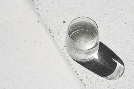 Glass with pure water on neutral concrete background with aesthetic sunlight shadow. Purity and...