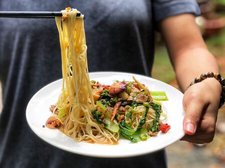 Fried spicy spaghetti with seafoods or Phad Kee Mao Spaghetti in white ceramic dish, hold by female hands.の写真素材