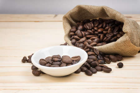 Coffee beans inside burlap bag and white bowl on wooden background. Selective Focus.