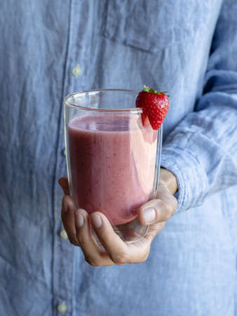 Female hand holding glass of pink smoothie. Cold strawberry-banana shake with coconut milk. Raw healthy drink with fruits and berries decorated strawberry. Vegan Smoothiesの写真素材