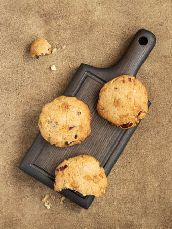 Oatmeal cookies with cranberry, cashew and white chocolate on a wooden tserving board. Brown background, small wooden bowl with nuts and dried berries. A bitten cookie with some crumbsの写真素材