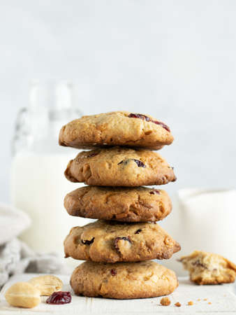 Stack of oatmeal cookies with cranberry, cashew nuts and white chocolate on a white wooden cutting board. Glass bottle of milk, kitchen towel and milk jug on the backgroundの写真素材