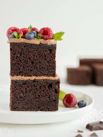 Two pieces of brownie cake on top of each other decorated chocolate cream cheese and raspberry, blueberry and mint leaves. White plate, white background, pieces of chocolate on theの写真素材