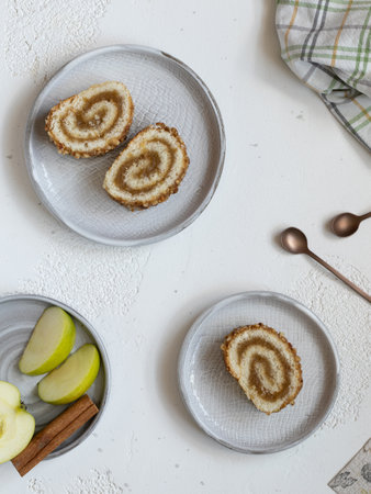 Pieces of biscuit roll with apple jam on a plates. Checkered kitchen, fresh green apple, cinnamon and teaspoons on white background, flat layの写真素材