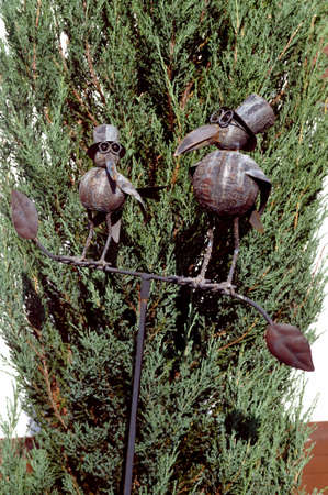 two metal puppet birds on a perch in front of a bush in frontviewの写真素材