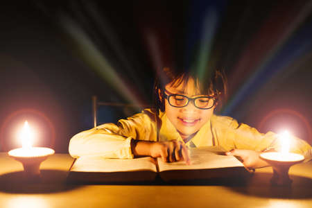 Cute little girl with glasses reading book with lighting candle at night time.の写真素材