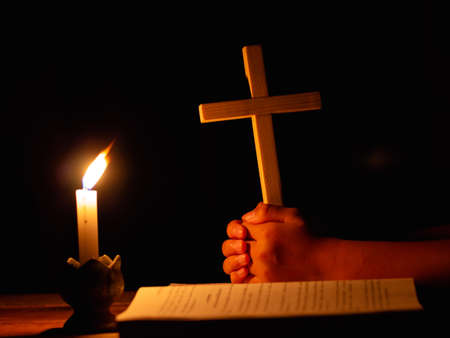 Human praying to the GOD while holding a crucifix symbol with lighting candle at night time.