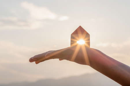 Hand holding wooden house toy against blue sunny sky with sun rays. solar power.
