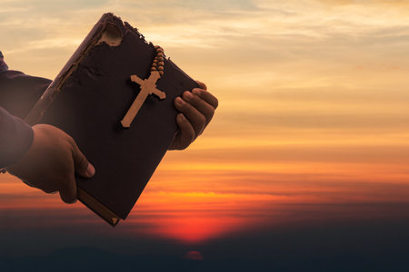 Hands holding Old Holy Bible book and Cross on sunset sky. Religion and Hope Concept.の写真素材