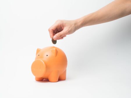 Woman Hand  putting coin into piggy bank for saving with pile of coins on white backgoound.A orange piggy bank are smile and happy .Many coin on the table.の写真素材