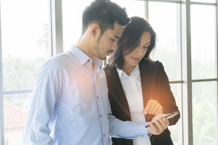 Business man and woman wearing suit, looking smartphone. Open space loft office. Panoramic windows background.の写真素材