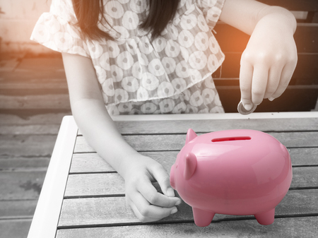 Little girl putting coin into piggy bank for saving with pile of coins on table at home.A orange piggy bank are happy.Chid put coin in piggy bang for saving for the future.の写真素材