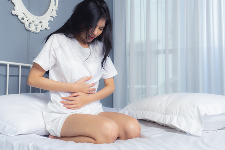 Young woman suffering from abdominal pain while sitting on white  bed at home. Do not oversleep againの写真素材