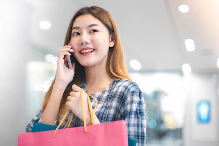Smiling young Asian woman with shopping colour bags over mall background. using a smart phone shopping online  and smiling while standing mall building. lifestyle conceptの写真素材