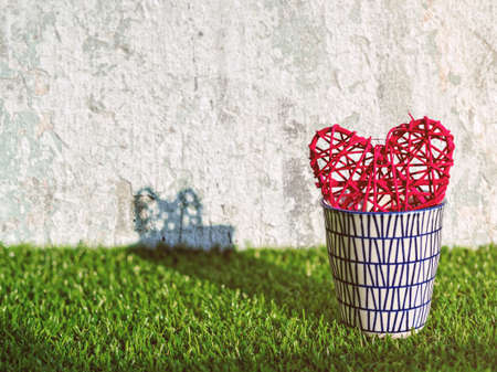 The wicker red heart is in a porcelain mug on the green grass. Behind there is a dilapidated wall.
