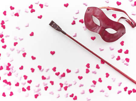 The Venetian mask and leather whip are on a light background with pink and red heart made of cloth. Romantic background for goods from a sex shop, costume party, Valentine's Dayの写真素材