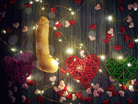 A flesh-colored dildo, three wicker hearts and a glowing garland are on a dark background. Around...