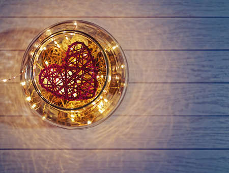 A woven red heart is on a light wooden background. Under the symbol of love are wicker balls of a different sizes and colors and a radiant garlandの写真素材