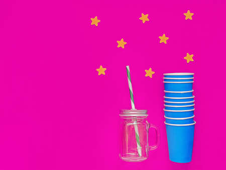 Empty blue paper cups are stacked. The distance is an empty glass jar with a handle, a lid with a hole and a straw. Purple background with yellow felt stars. Idea for a party, celebrating somethingの写真素材