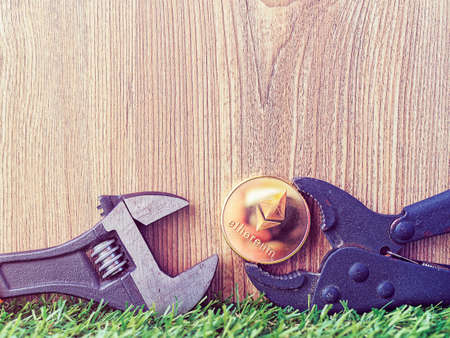 The coin of the crypto currency Ethereum is clamped in a wrench. The objects are on the green grass....